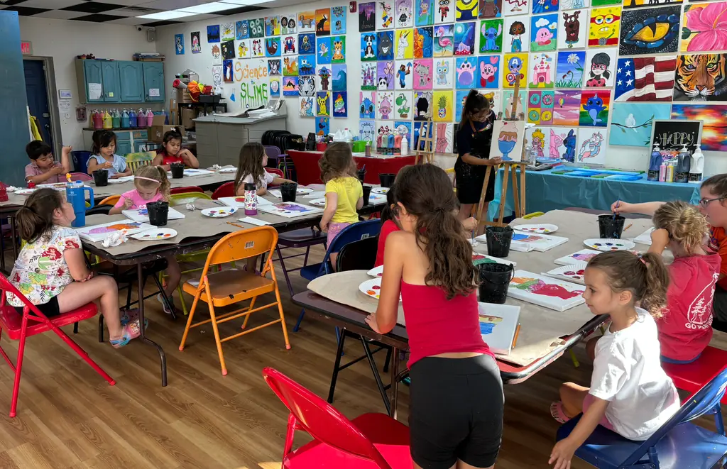 Kids painting at long tables in the studio, under a wall covered in colorful student canvases